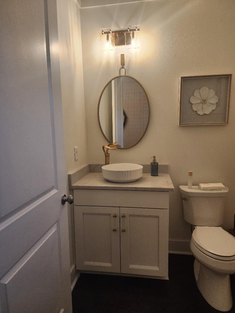 Modern bathroom with cream vanity, round mirror, vessel sink, toilet, wall art, and dark tile flooring