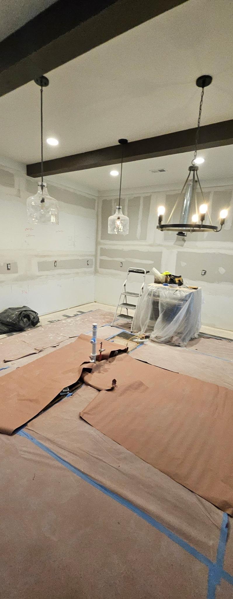 Interior renovation in progress showing unfinished drywall, pendant light fixtures hanging from dark beam, and protective tarps covering the floor