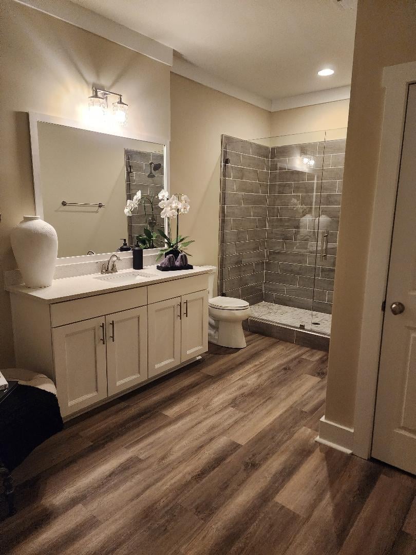 Modern bathroom with white vanity, large mirror, gray tile shower enclosure, toilet, and wood-look flooring