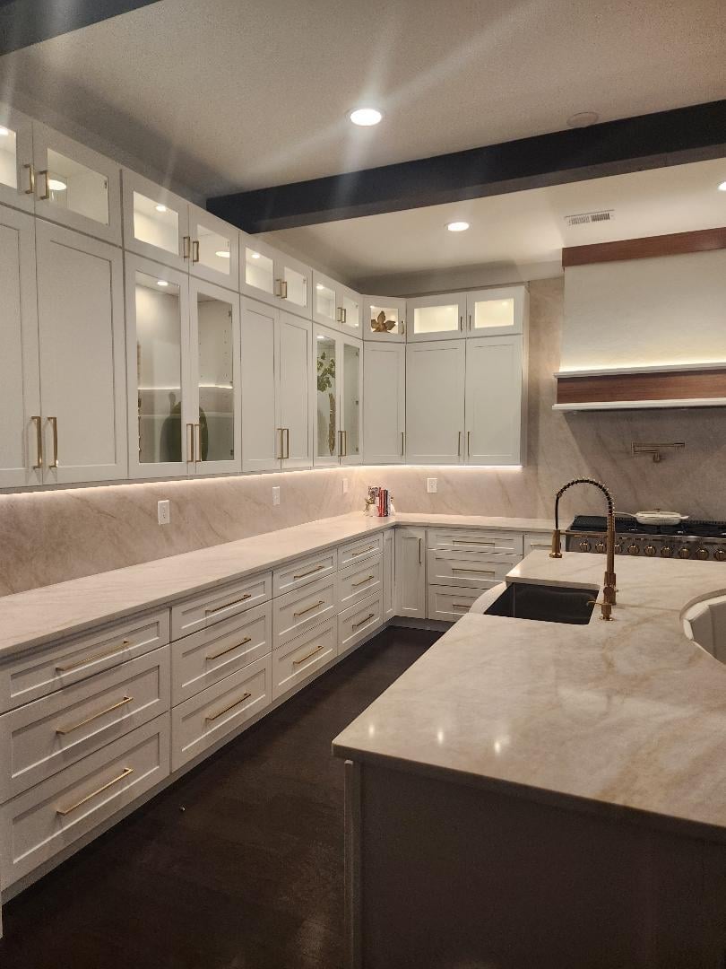 Modern white kitchen with marble countertops, white cabinetry, dark island, and stainless steel sink under recessed lighting