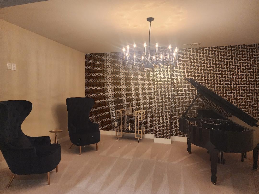 Elegant music room with grand piano, two black wingback chairs, decorative candelabra, and textured brick accent wall under chandelier lighting